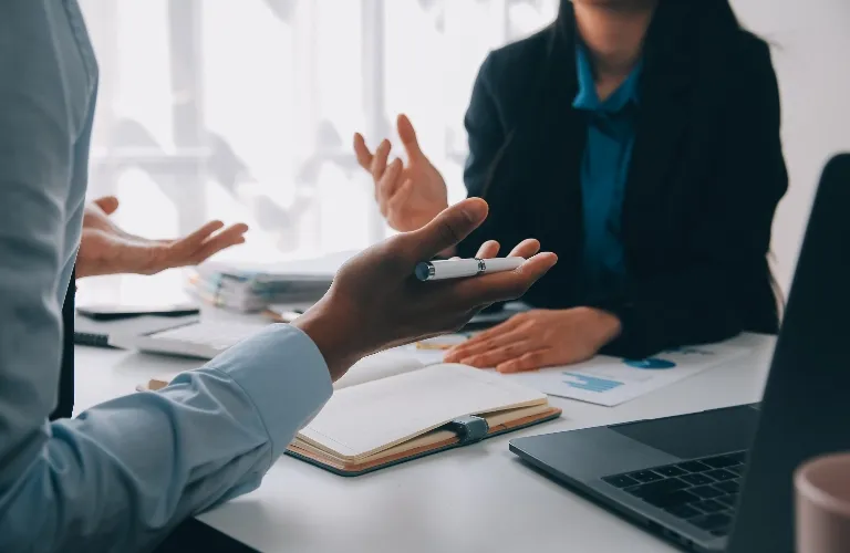 Two professionals engaged in discussion at a desk with a laptop, notebook, and documents.