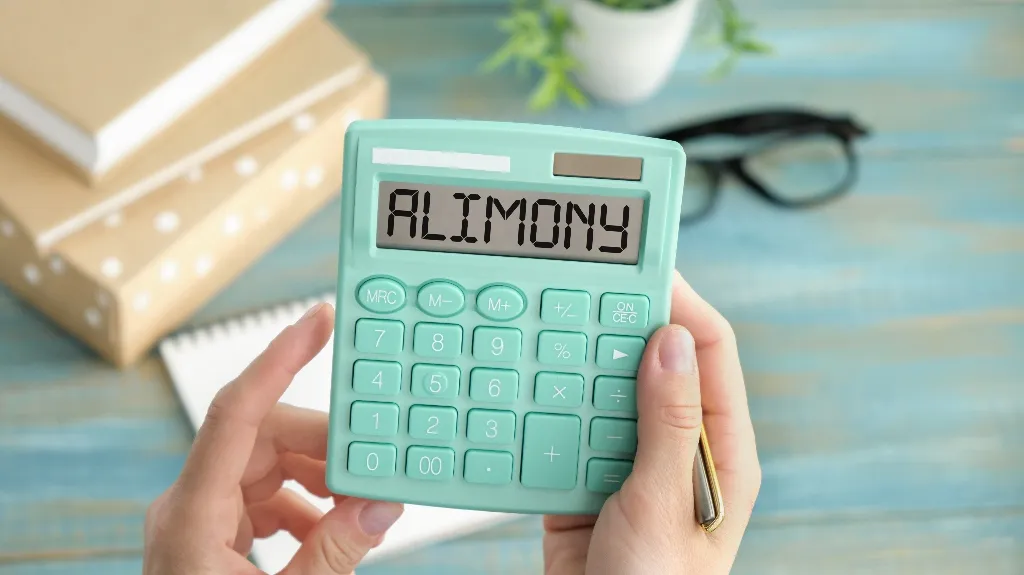 Hands holding a mint green calculator displaying the word "ALIMONY" on a blue wooden surface.