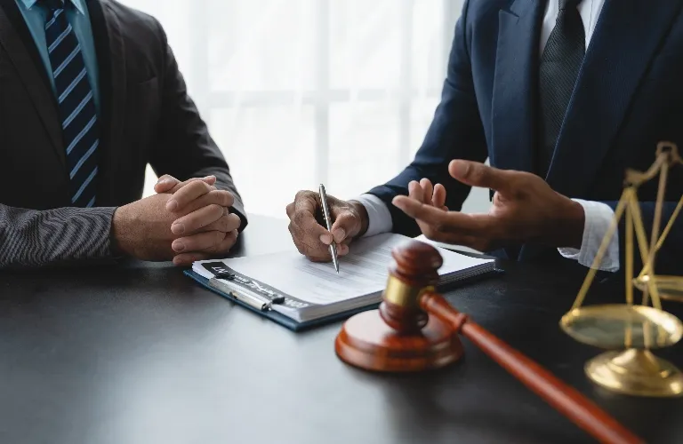 Two men in suits discussing legal documents with a gavel and scales of justice on the desk.