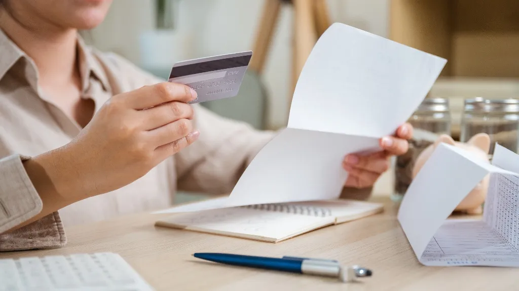 Person holding a credit card and a document, with a pen and notebook on a wooden desk.