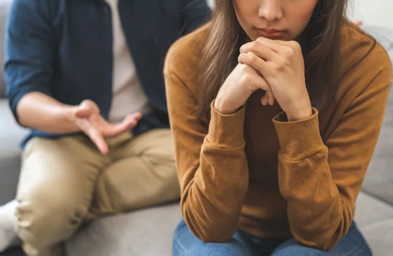 Two people seated on a couch, woman in brown sweater appears distressed, man in blue shirt gesturing.