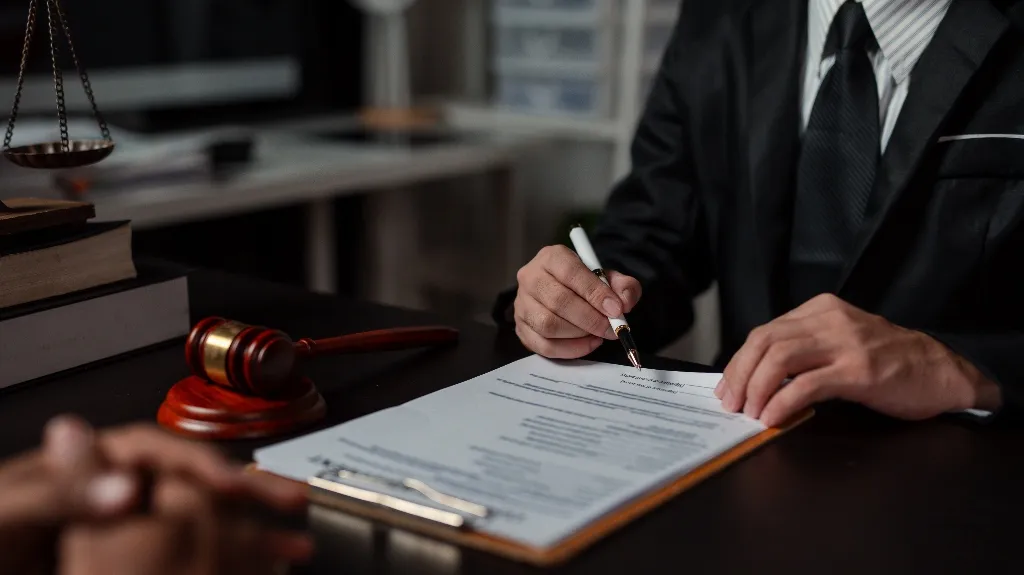 Man in black suit signing legal documents on clipboard, with judge's gavel and scales on desk.