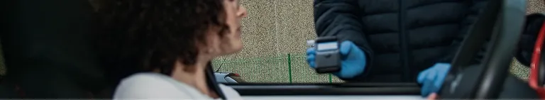 Person in black jacket and blue gloves holding a device near a seated woman in a car.