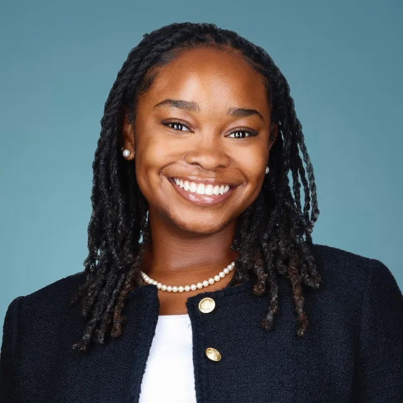 Professional portrait of a smiling woman with dreadlocks, pearl necklace, and navy blazer.