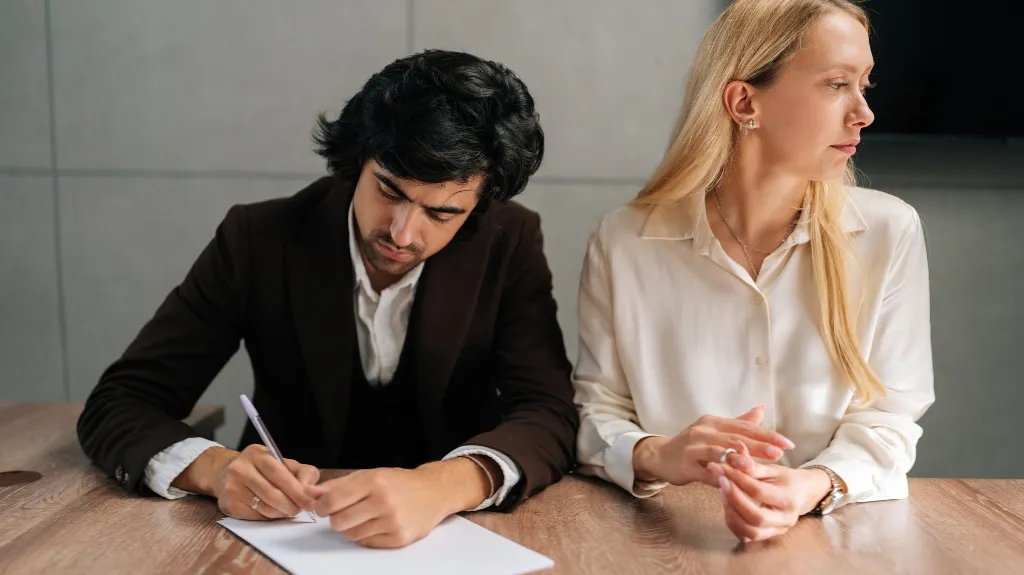 Two professionals seated at a wooden table; man writing on paper, woman looking away.