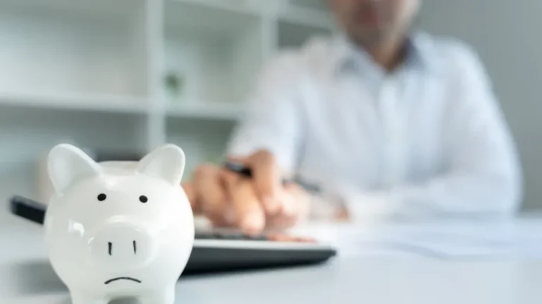 White ceramic piggy bank in foreground with person using calculator in background.