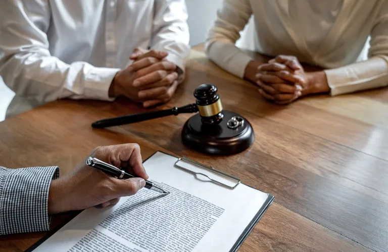 Two individuals sit across a wooden table from a person holding a pen and a legal document, with a judge's gavel placed on the table.