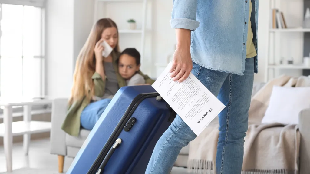 Person in jeans holding a divorce decree and suitcase, with distressed woman and child in background.