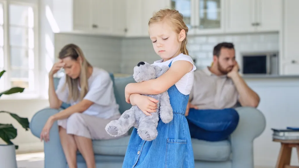 Young girl in blue dress holding a stuffed animal, parents sitting upset on couch in background.