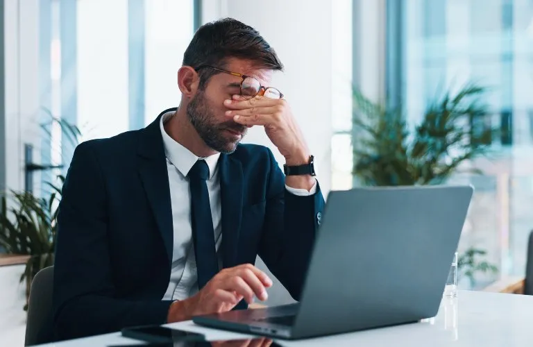 Professional man in dark suit and glasses sitting at desk, rubbing eyes in front of laptop.