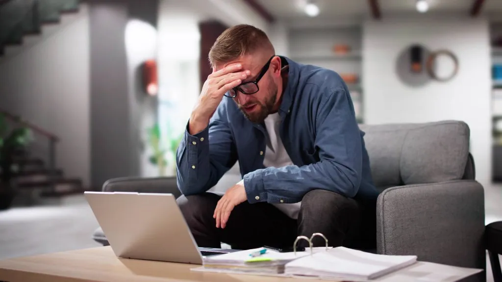 Man wearing glasses and a blue shirt sitting on a gray sofa, stressed while looking at a laptop.