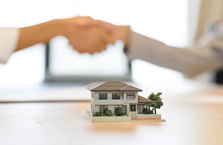 Two people shaking hands behind a detailed miniature house model on a wooden table.