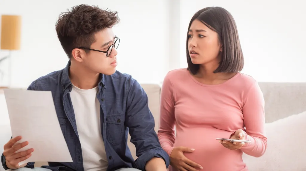 A concerned young pregnant woman holding a phone talks to a man holding a document.