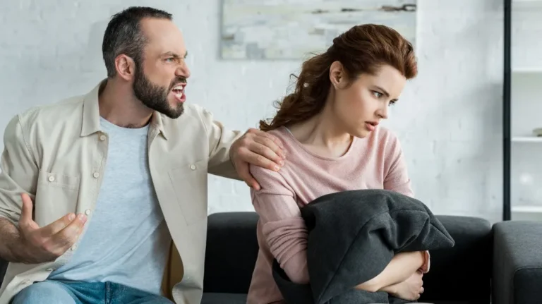Man angrily confronting woman on sofa, woman looking away holding black pillow.
