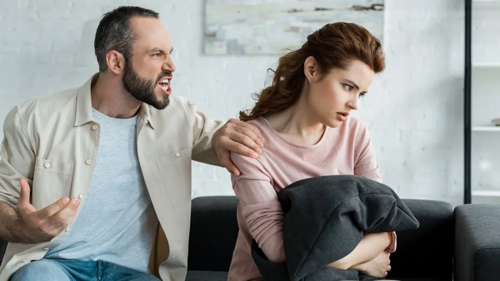Man angrily confronting woman on sofa, woman looking away holding black pillow.
