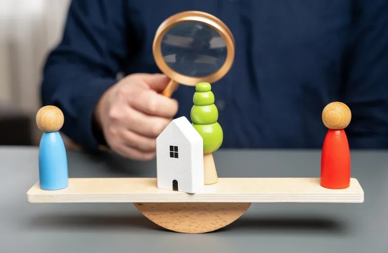 Wooden figurines, house, and tree balanced on a seesaw with a hand holding a magnifying glass in the background.