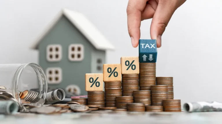 Hand placing a blue block labeled "TAX" on stacked coins with percentage blocks and a small house model in the background.