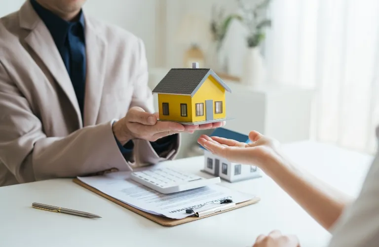Person in beige suit presenting yellow house model to another person over documents and calculator on white table.