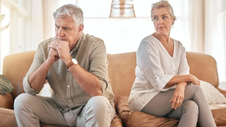 Mature man and woman sitting on a beige couch, appearing distant and contemplative.