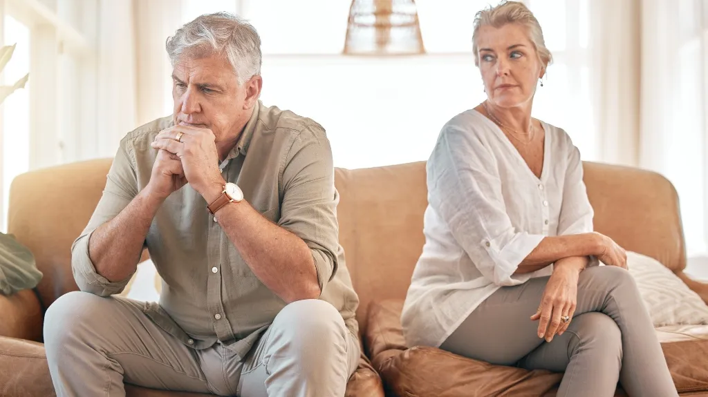 Mature man and woman sitting on a beige couch, appearing distant and contemplative.