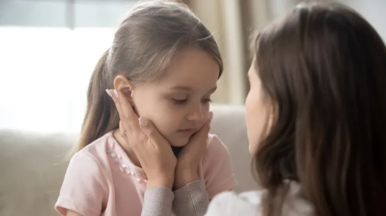 Adult woman gently holding young girl's face, both with brown hair, indoors.